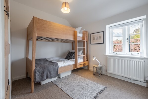 Un lit superposé avec un bureau et une chaise, un radiateur, une fenêtre donnant sur des immeubles et un tableau encadré accroché au mur.