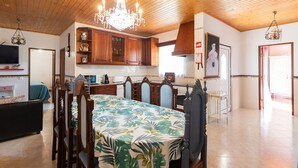 A spacious room with a wooden ceiling, a dining table with chairs, a chandelier, and a kitchen area with wooden cabinets and a tiled backsplash.