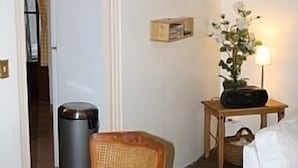 Une pièce comprenant une table à manger en bois, une chaise en osier, une petite table d’appoint en bois avec une lampe, une poubelle et un lit blanc.