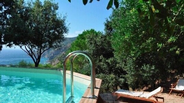 A clear swimming pool with a wooden deck and lounge chairs, surrounded by lush greenery and overlooking a mountainous landscape.