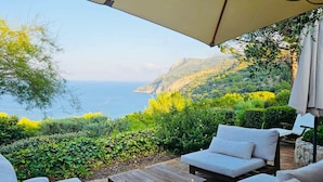 An outdoor seating area with white cushioned sofas and a wooden table, overlooking a scenic coastal landscape with lush greenery and mountains.