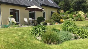 A house with a sloped roof, a patio with chairs and an umbrella, and a well-maintained garden with various plants and shrubs.