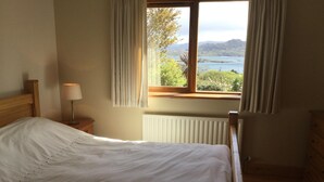 A bedroom with a bed, a nightstand with a lamp, a window with curtains, and a view of a lake and mountains.