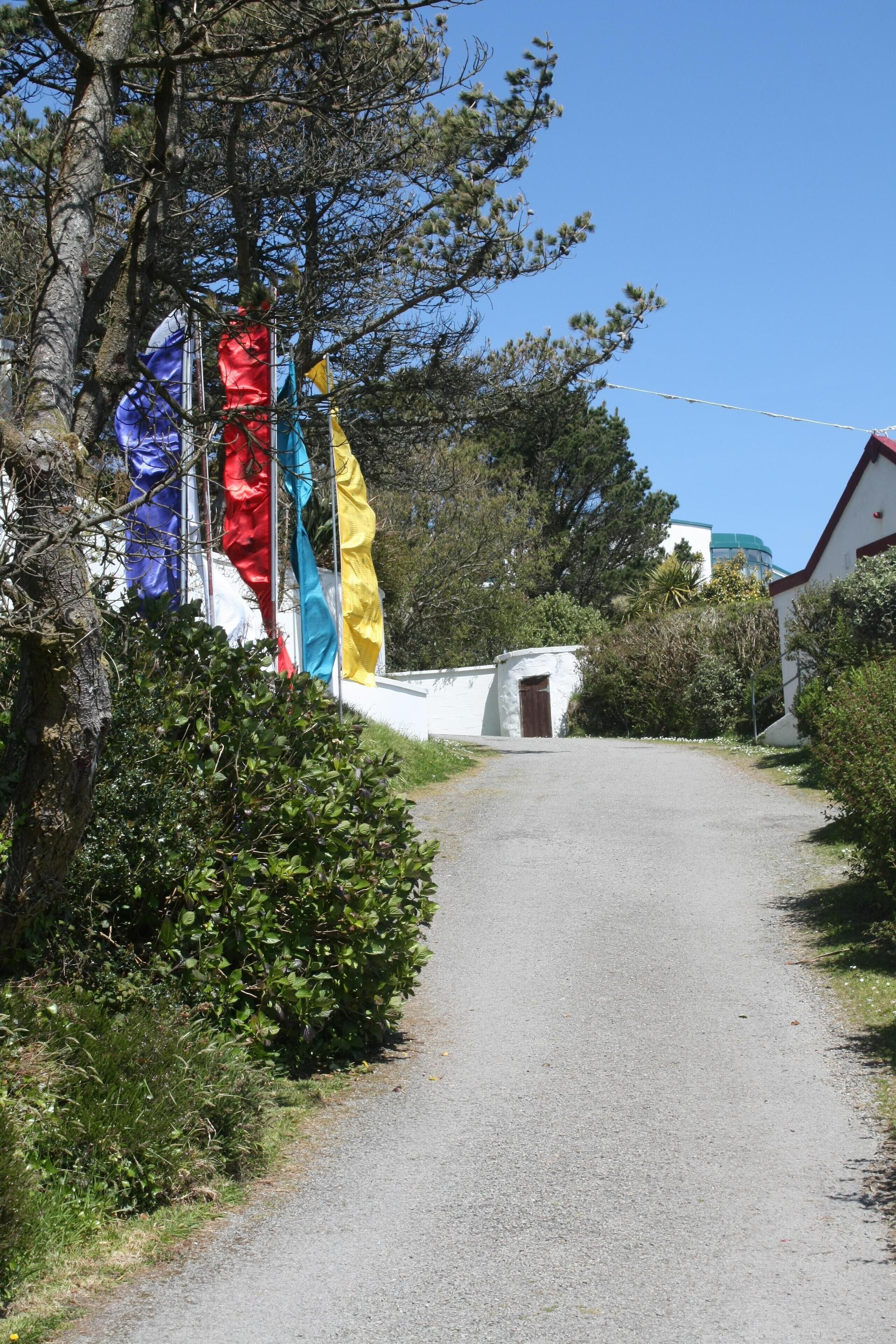 Ein Kiesweg führt zu einem weißen Gebäude mit rotem Dach, umgeben von Grün und mehreren bunten Flaggen.