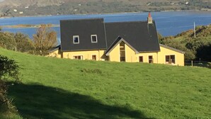 A house with a dark roof and yellow walls, situated on a grassy hill overlooking a body of water and mountains.