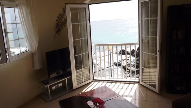 A living room with a TV, a table with a red object, and a balcony with a view of the sea.