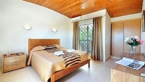 Un dormitorio con techo de madera, una cama con ropa de cama a rayas, una mesita de noche, un escritorio con un jarrón de flores y una ventana con cortinas.