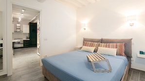 Un dormitorio con cama, almohadas, una mesita de noche y un mueble auxiliar. Se ve una cocina a través de una puerta abierta.