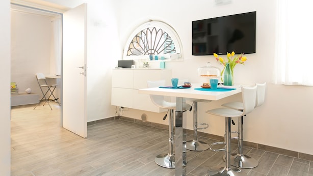 Una cocina moderna con encimera blanca, dos taburetes de bar, un televisor montado en la pared y un ventanal con un espejo decorativo.