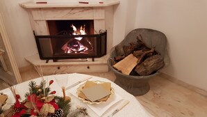 A dining table set with a white tablecloth, a glass of wine, cutlery, and a centerpiece with pinecones and greenery. A fireplace with burning logs is visible in the background, and a stone chair with logs is to the right. - Villa "LA GARITTA" Two steps from the magical village (Finale Ligure)