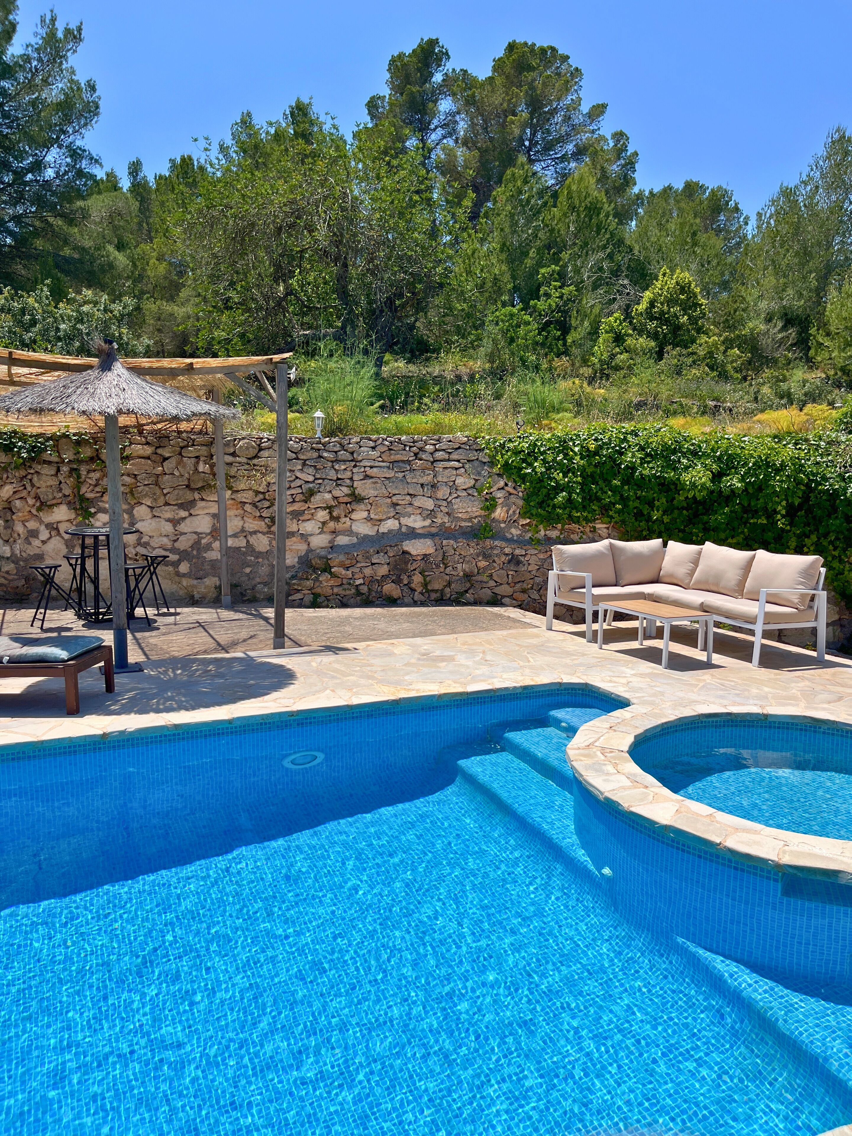 Una piscina con un muro de piedra y una pérgola de madera. Hay un sofá color beige y una mesa con sillas.