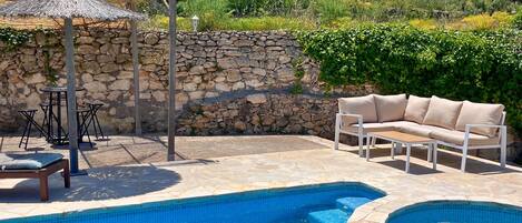 Una piscina con un muro de piedra y una pérgola de madera. Hay un sofá color beige y una mesa con sillas.