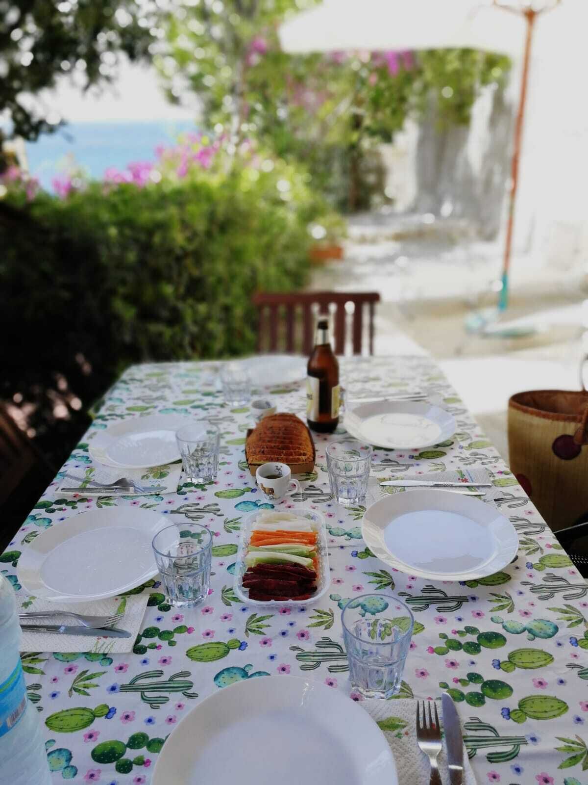 Mesa dispuesta para una comida con platos, vasos, cubiertos, un pan y una botella, al aire libre con vistas a vegetación y un edificio al fondo.