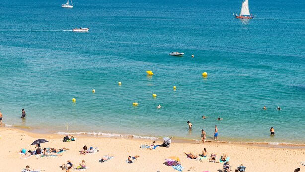 A beach with people sunbathing and swimming, boats on the water, and a clear sky.