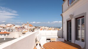 A rooftop terrace with a wooden table and white seating, overlooking a cityscape with buildings and a church.