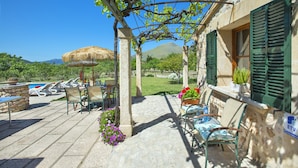 Une terrasse avec un sol en pierre, des meubles d’extérieur, une pergola couverte de plantes grimpantes, un parasol en chaume et une piscine en arrière-plan.
