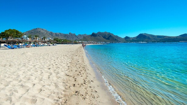 Une plage avec une eau d’un bleu limpide, un rivage sablonneux et des montagnes en arrière-plan.