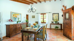 Un coin repas avec une table en bois dressée pour quatre personnes, entourée de chaises en osier. Un lustre est suspendu au plafond, et on peut voir deux portes, dont l’une est ornée d’un motif décoratif en métal.