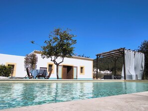 Un espace aménagé au bord de la piscine, avec un bâtiment blanc, un arbre et un coin salon abrité.