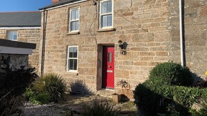 A stone house with a red door, two windows, and a small garden area with plants and a gravel path.