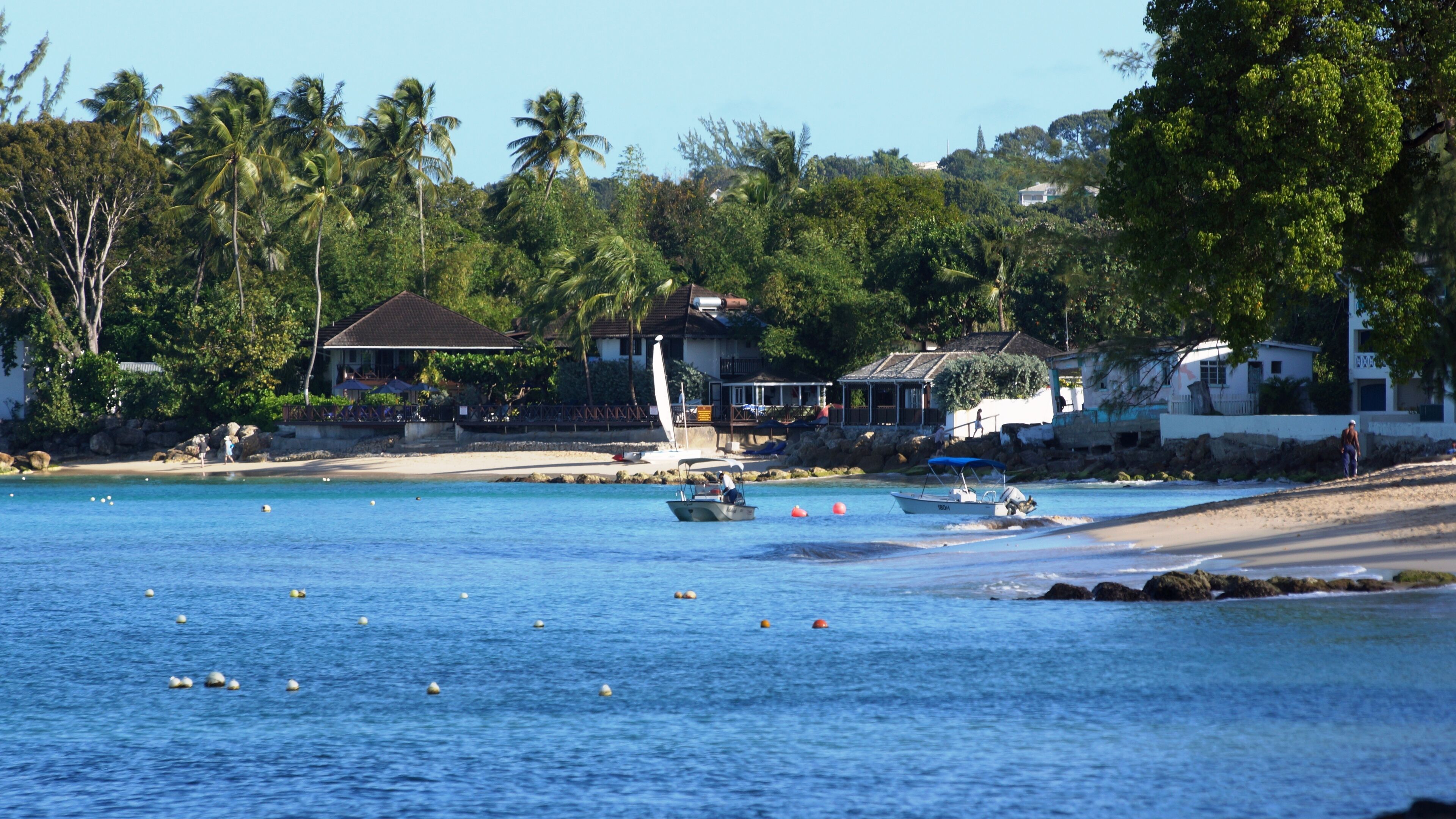 Une zone côtière avec une eau bleue et limpide, des bateaux et une plage de sable. On aperçoit des maisons et des arbres au loin.