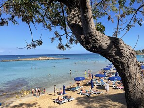 Een strand met helderblauw water, mensen die zwemmen en zonnebaden, en een rotsachtige landtong in de verte.