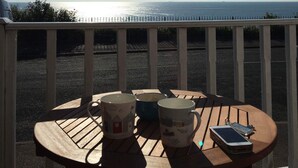 A wooden table with three mugs and a smartphone on a balcony overlooking a beach.