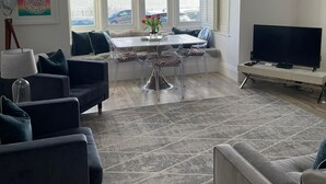 A modern living room with a dining area, large windows, a flat-screen TV, and a view of the sea.