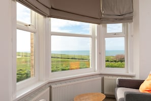 A room with a bay window overlooking a coastal landscape, a radiator, and a seating area with a cushion. - Stunning seaside with cinema room, sea views  - ideal for friends and families (Newquay)