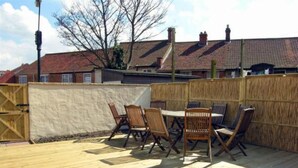 A wooden deck with a table and chairs, surrounded by a wooden fence, with houses in the background.