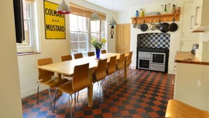 A dining area with a checkered floor, a wooden table, and chairs. There is a kitchen with a checkered backsplash and hanging pots.