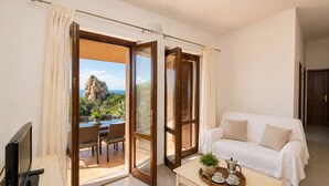 A room with a white sofa, a dining table with chairs, a TV, and a view of a rocky landscape through open doors.