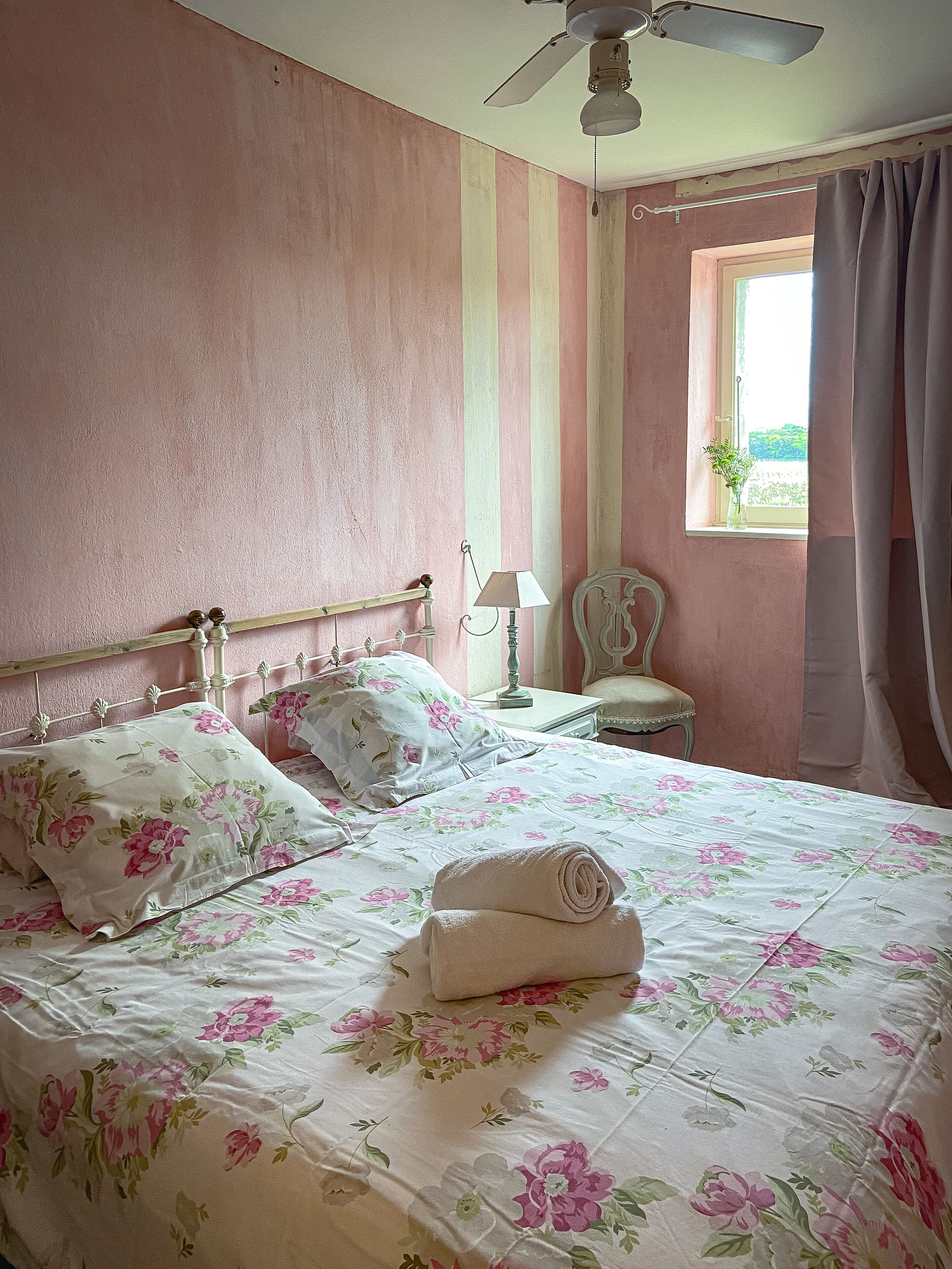 A neatly made bed with floral bedding, a bedside table with a lamp, and a chair in a room with pink walls and a window.
