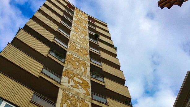 Um edifício alto com um design vertical marcante, apresentando um mural grande em uma das laterais e várias sacadas.