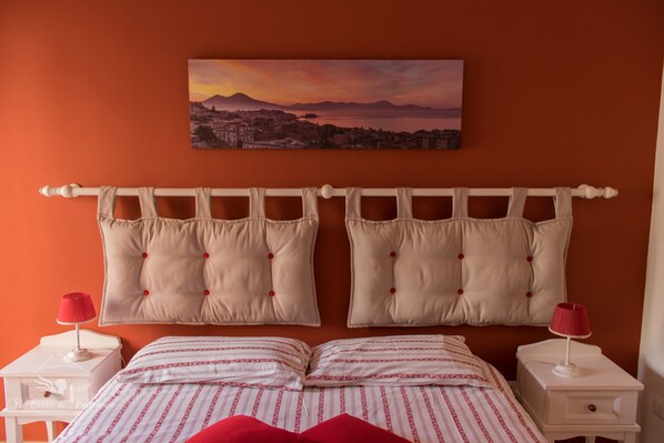 A bedroom with two beds, a cityscape painting, and a red lamp on each nightstand.