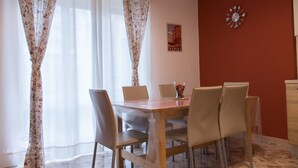 Uma sala de jantar com uma mesa de madeira e seis cadeiras, uma cortina florida, um relógio de parede e um quadro emoldurado.