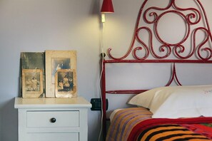 A bedroom with a white nightstand, two vintage photographs, a red metal headboard, and a striped bedspread. - Vico Muccio - historical house, finely restructured with a splendid view on the historical Baroque city of Modica (Modica)