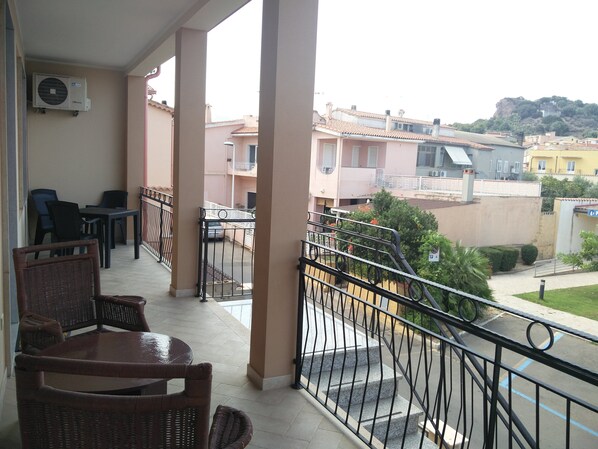 A balcony with wicker furniture, a table, and chairs, overlooking a residential area with buildings and a hill in the background.