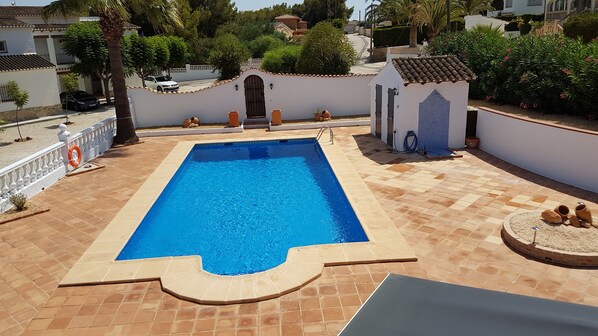 Une piscine rectangulaire avec une terrasse carrelée, entourée d’un mur blanc surmonté d’un toit en tuiles, et un petit bâtiment blanc avec un toit en tuiles situé à droite.