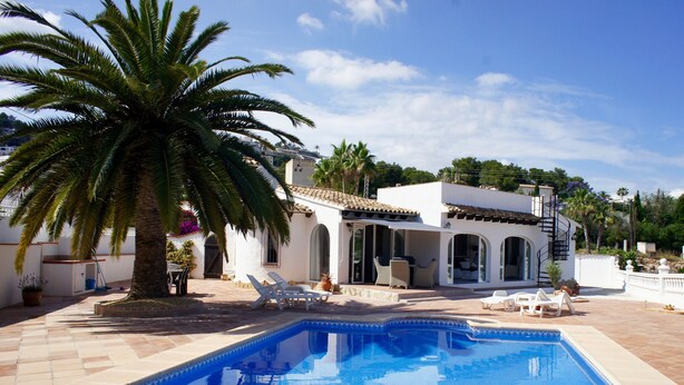 Zona de piscina con una piscina azul cristalina, edificio blanco con ventanas arqueadas y una palmera.