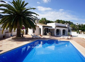 Un espace aménagé au bord d’une piscine à l’eau d’un bleu limpide, un bâtiment blanc doté de fenêtres en arc de cercle et un palmier.