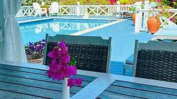 A poolside table with a white vase of purple flowers, white chairs, and a view of a pool with palm trees and a white fence.