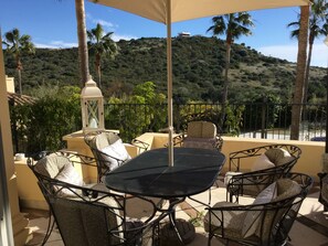 A terrace with outdoor furniture, a table, and chairs, overlooking a hillside with vegetation and a building in the distance.