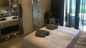 Une chambre d’hôtel avec un grand lit, un coin salon avec un fauteuil, un miroir et un lustre.