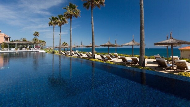 Un espace aménagé au bord de la piscine, avec des chaises longues et des palmiers, offrant une vue sur une mer d’un bleu limpide.