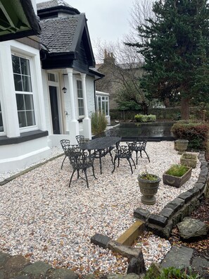 A white building with a black roof, a gravel patio with a table and chairs, and a stone wall with planters. - Extended Victorian Manor House With Conservatory / large Garden, sleeps 10 (Cardross)