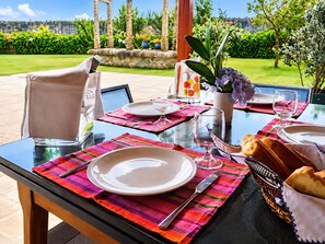 An outdoor dining area with a table set for two, featuring a glass top, colorful placemats, white plates, and a basket of bread.