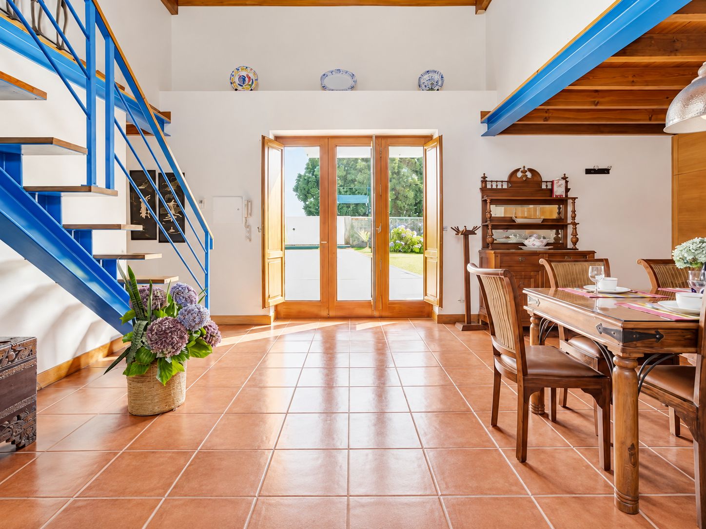A spacious room with a wooden staircase, a dining area with a table and chairs, and a large sliding glass door leading to an outdoor area.