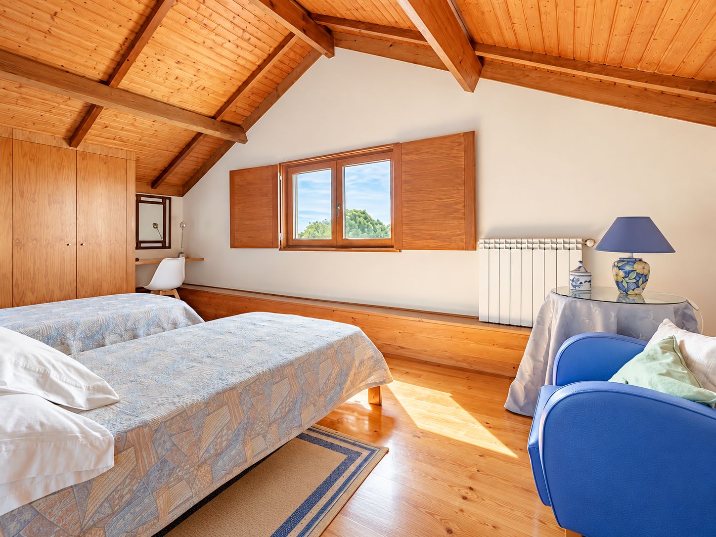 A bedroom with two beds, a window, a radiator, and a table with a lamp.