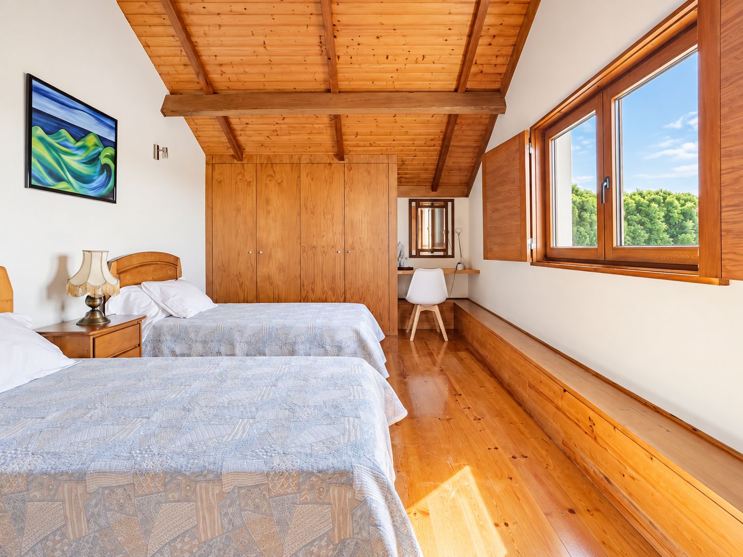 A room with two beds, a wooden ceiling, a window with a view of greenery, and a painting on the wall.